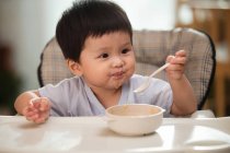 Adorable asiático niño sosteniendo cuchara y mirando lejos mientras comer en casa - foto de stock