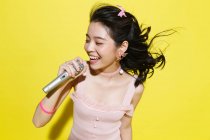 Beautiful happy young asian woman holding microphone and singing on yellow background — Stock Photo