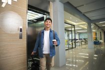 Красивый счастливый молодой азиатский бизнесмен держит кофе, чтобы пойти и тянуть самобалансирующийся скутер возле лифта — стоковое фото