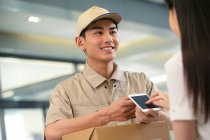 Cropped shot of smiling young courier with cardboard box looking at businesswoman using smartphone in office — Stock Photo