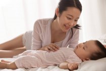 Красивая улыбающаяся молодая азиатская женщина, глядя на своего очаровательного ребенка, спящего на кровати — стоковое фото