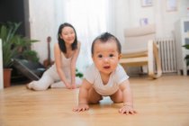 Smiling young mother looking at beautiful baby crawling on floor at home — Stock Photo