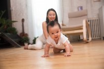 Glückliche junge Mutter blickt auf entzückendes Baby, das auf dem Boden kriecht und in die Kamera lächelt — Stockfoto