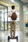 Back view of young businessman with backpack riding self-balancing scooter in office — Stock Photo
