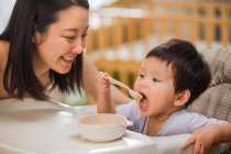 Feliz jovem asiático mãe olhando adorável bebê segurando colher e comer em casa — Fotografia de Stock