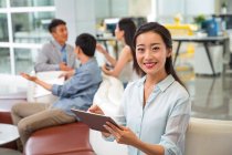 Bella giovane donna d'affari asiatica utilizzando tablet digitale e sorridente alla macchina fotografica, colleghi che parlano dietro in ufficio — Foto stock