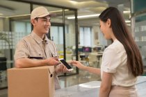 Sourire jeune asiatique courier avec boîte en carton regardant femme d'affaires en utilisant smartphone dans le bureau — Photo de stock