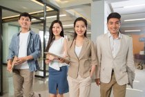 Молоді професійні азіатські ділові люди, що стоять з кишенями і посміхаються на камеру в офісі — стокове фото