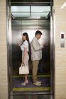 Side view of young asian businessman and businesswoman standing back to back and using smartphones in elevator — Stock Photo
