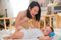 Felice giovane madre guardando adorabile bambino sdraiato nella culla e giocando con i giocattoli in gomma — Foto stock