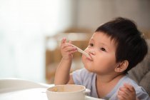 Adorable asian toddler eating with spoon and looking up at home — Stock Photo