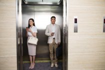 Yougn asiatico businessman holding caffè a andare e businesswoman utilizzando smartphone mentre standing insieme in ascensore — Foto stock