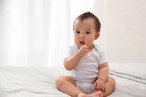 Вид спереду чарівного азіатського немовляти тримає шматок очищених фруктів і дивиться на камеру, сидячи на ліжку — стокове фото