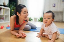 Feliz joven madre acostado en yoga mat y mirando adorable bebé en casa - foto de stock