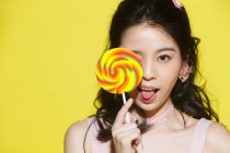 Beautiful young asian woman holding colorful lollipop and showing tongue out isolated on yellow background — Stock Photo