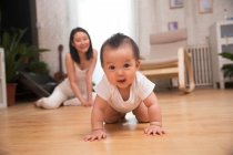 Прелестный счастливый азиатский ребенок ползает по полу и смотрит в камеру, улыбаясь молодая мама сидит позади дома — стоковое фото