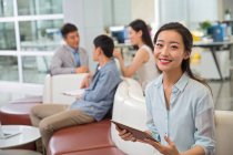 Schöne junge asiatische Geschäftsfrau hält ein digitales Tablet in der Hand und lächelt in die Kamera, während Kollegen im Büro hinterherarbeiten — Stockfoto
