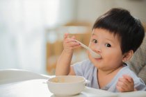 Adorable asian toddler child holding spoon and eating at home — Stock Photo