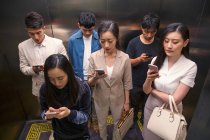 Высокий угол обзора молодых азиатских людей, стоящих в лифте и использующих смартфоны — стоковое фото
