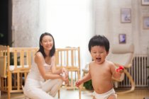 Glückliche junge Mutter schaut entzückend aufgeregtes Kleinkind an, das rotes Spielzeug in der Hand hält und zu Hause spaziert — Stockfoto