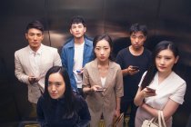 Vue grand angle de jeunes asiatiques sérieux tenant des smartphones et regardant la caméra dans l'ascenseur — Photo de stock