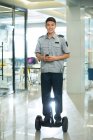 Lächelnder junger asiatischer Wachmann, der selbstbalancierende Roller fährt und im Büro Walkie-Talkie benutzt — Stockfoto