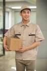Красивый улыбающийся молодой азиатский курьер, держащий картонную коробку в офисе — стоковое фото