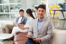 Красивый молодой азиатский бизнесмен, использующий цифровой планшет и улыбающийся на камеру в офисе — стоковое фото