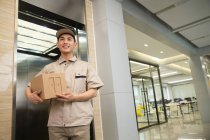 Faible angle de vue de sourire jeune asiatique livraison homme tenant boîte en carton dans le centre d'affaires — Photo de stock