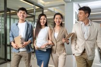 Glückliche junge professionelle asiatische Geschäftsleute halten Klemmbretter in der Hand und lächeln in die Kamera, während sie gemeinsam im Büro spazieren gehen — Stockfoto