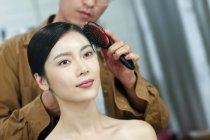 Recortado disparo de hombre sosteniendo cepillo de pelo y haciendo peinado hermosa joven asiática mujer en salón de belleza - foto de stock