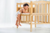Вид в полный рост красивого малыша в пеленках, залезающего на детскую кроватку и отводящего взгляд от дома — стоковое фото