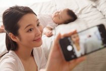 Счастливая молодая мать делает селфи со смартфоном, пока ребенок спит на кровати — стоковое фото
