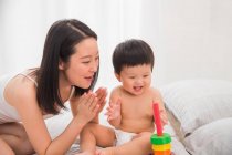 Happy young mother looking at adorable baby playing with colorful toy on bed — Stock Photo