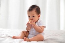 Full length view of adorable asian infant holding baby bottle with water and sitting on bed — Stock Photo