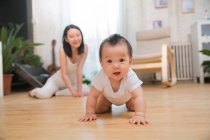 Adorable asiático bebé arrastrándose en suelo y sonriendo a cámara mientras feliz madre sentado detrás en casa - foto de stock