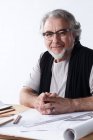 Professionelle reife Architekt in Brille sitzt am Schreibtisch mit Entwürfen und lächelt in die Kamera — Stockfoto