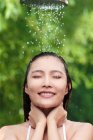 Приваблива усміхнена молода азіатська жінка з закритими очима приймає душ на зеленому природному фоні — стокове фото