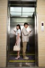 Вид сбоку молодого бизнесмена и бизнесвумен, стоящих спина к спине и пользующихся смартфонами в лифте — стоковое фото