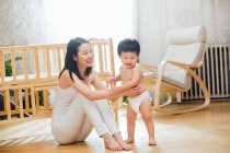Glückliche junge asiatische Mutter schaut Kleinkind in Windel an, das zu Hause zu gehen beginnt — Stockfoto