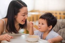 Feliz jovem asiático mulher olhando para bonito pouco criança segurando colher e comer em casa — Fotografia de Stock