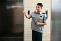 Sonriente joven asiático guardia de seguridad celebración walkie-talkie y apuntando lejos cerca de ascensores - foto de stock