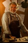 Professionnel mature mâle créateur de bijoux dans tablier debout sur le lieu de travail et souriant à la caméra — Photo de stock