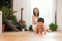 Felice giovane madre guardando il suo adorabile bambino eccitato strisciare sul pavimento a casa — Foto stock