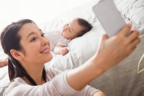 Selective focus of young mother taking selfie with smartphone while baby sleeping on bed — Stock Photo
