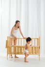 Улыбающаяся молодая мама смотрит на милого малыша в подгузнике, стоящего рядом с кроваткой дома — стоковое фото