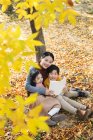 Alto ángulo vista de sonriente asiático madre lectura libro a hija e hijo cerca de árbol en otoñal parque - foto de stock