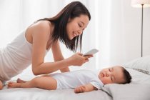 Усміхнена молода азіатка тримає смартфон і фотографує чарівну дитину, яка спить на ліжку — стокове фото