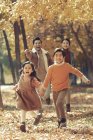 Felices padres jóvenes y lindos niños corriendo en el bosque de otoño y sonriendo a la cámara - foto de stock