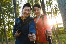 Счастливая молодая азиатская пара с биноклем и треккинговыми палочками, улыбающаяся в лесу перед камерой — стоковое фото